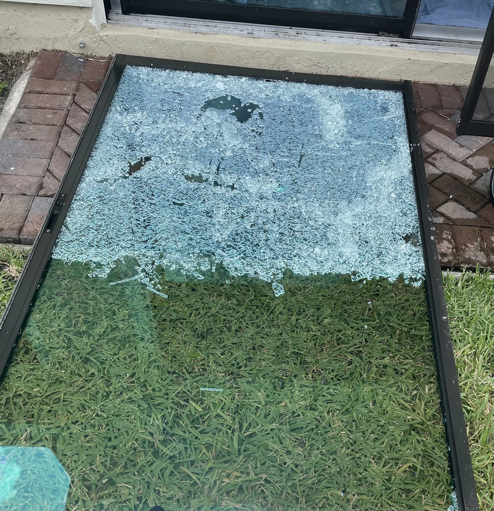Shattered tempered glass sliding door panel lying flat on a lawn beside a brick paver patio
